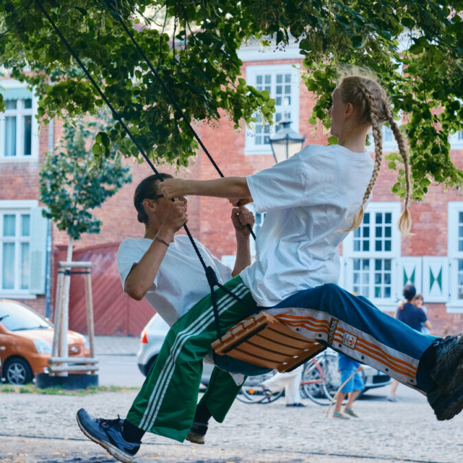 Ein Foto von der Aufführung des Tanzstücks "Balanç", bei dem zwei junge Menschen auf einer Schaukel schwingen. Im Hintergrund sind Häuser des Holländischen Viertels in Potsdam zu sehen.