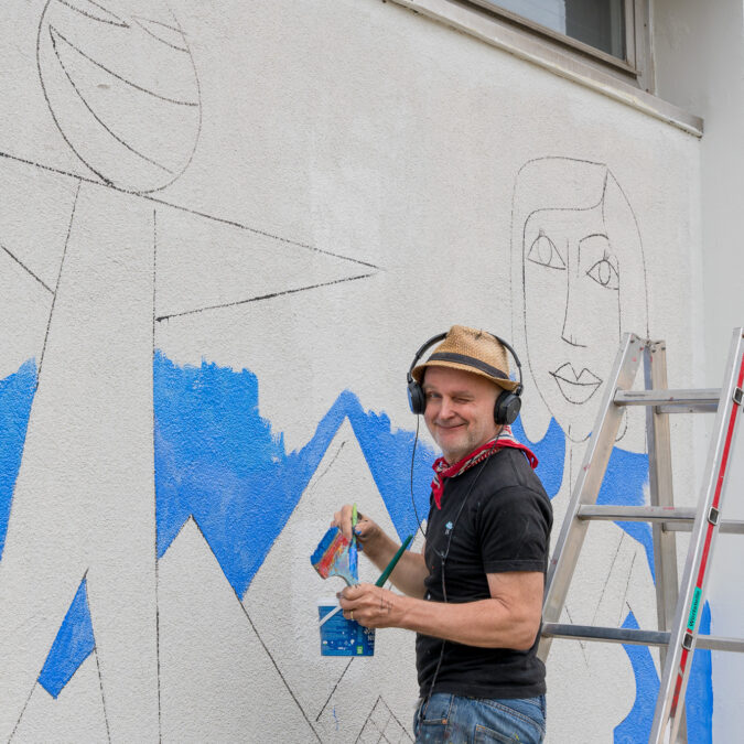 Ein Foto davon, wie Jim Avignon eine blaue Hintergrundfläche seines Murals an das RZ malt.