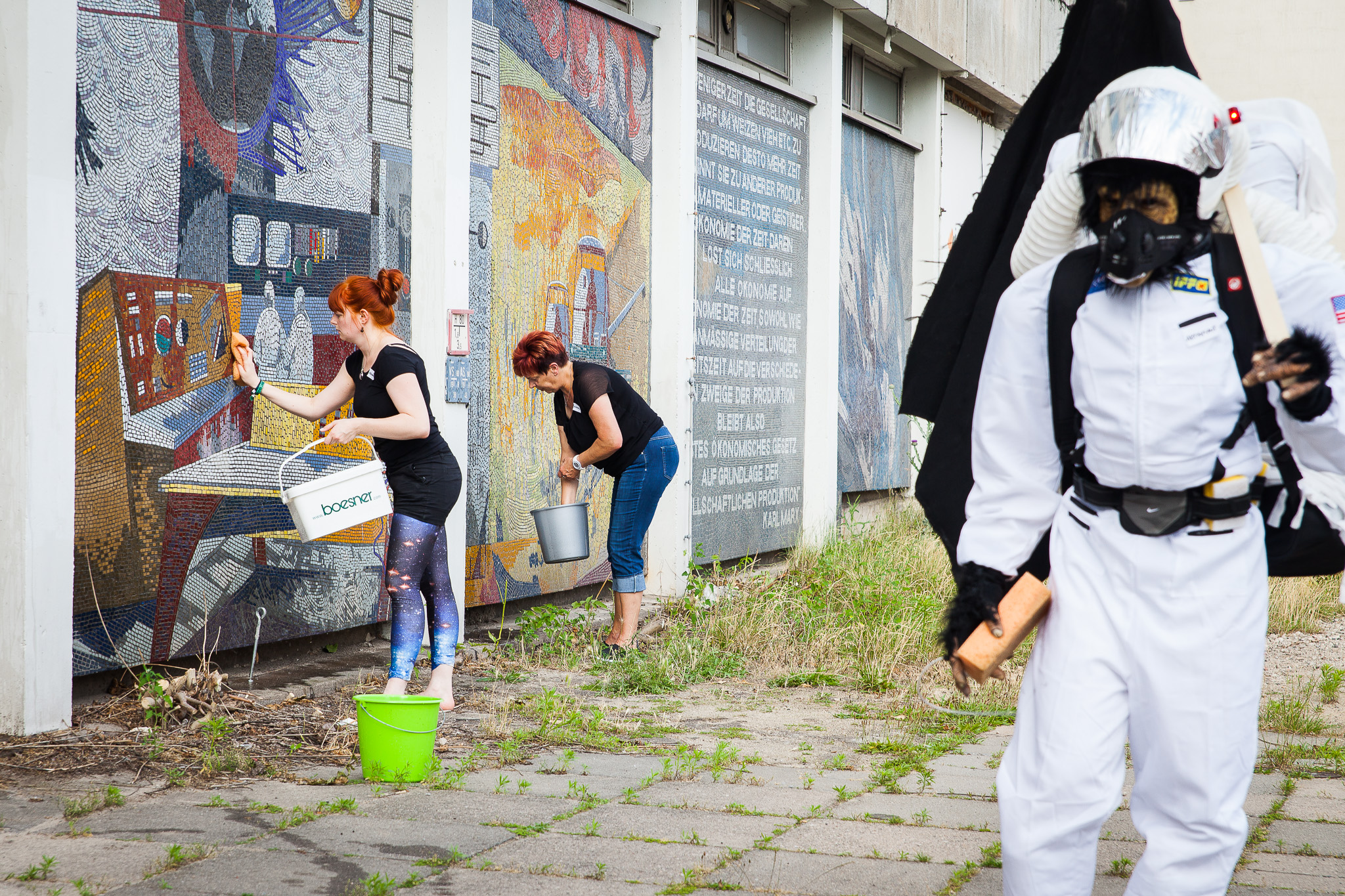 Foto der Mosaikwaschungsaktion am Rechenzentrum: links waschen zwei Frauen die Mosaiktafeln ab, rechts ist eine Person mit Affen-Kosmonauten-Kostüm zu sehen