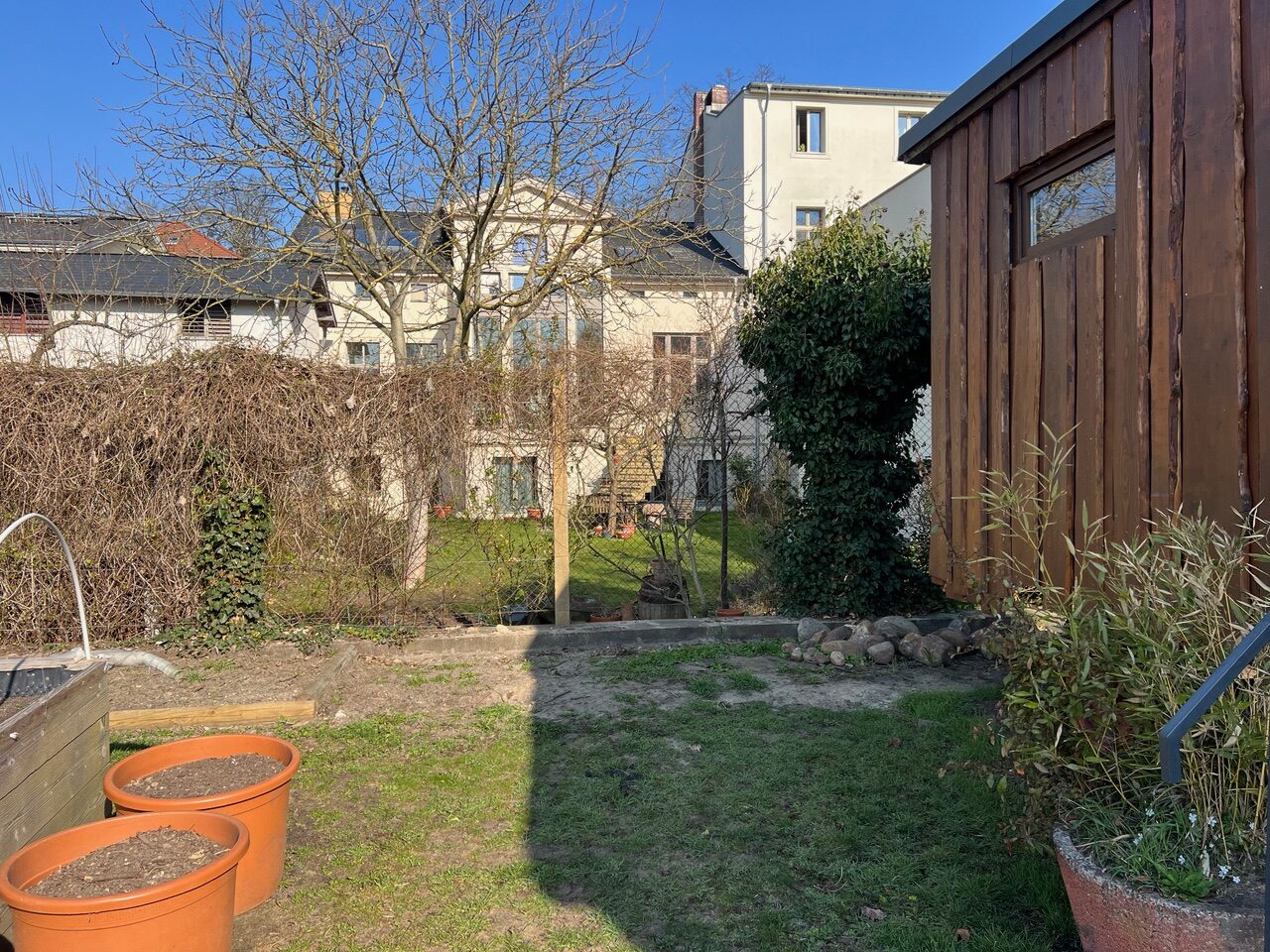 Eine Ansicht des "Essbaren Stadtgartens" an der Schopenhauerstraße: Man sieht rechts ein hölzernes Gebäude und links Blumentöpfe, in der Mitte Rasen.