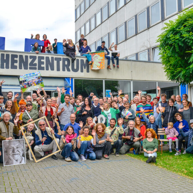 Ein farbenfrohes Foto von einer Gruppe Menschen, die vor dem Eingang des Rechenzentrums posieren. Einige sitzen oder stehen auf dem Dach des Eingangsgebäudes. Manche Menschen haben Utensilien dabei, die auf ihren Beruf oder ihr Hobby hindeuten.