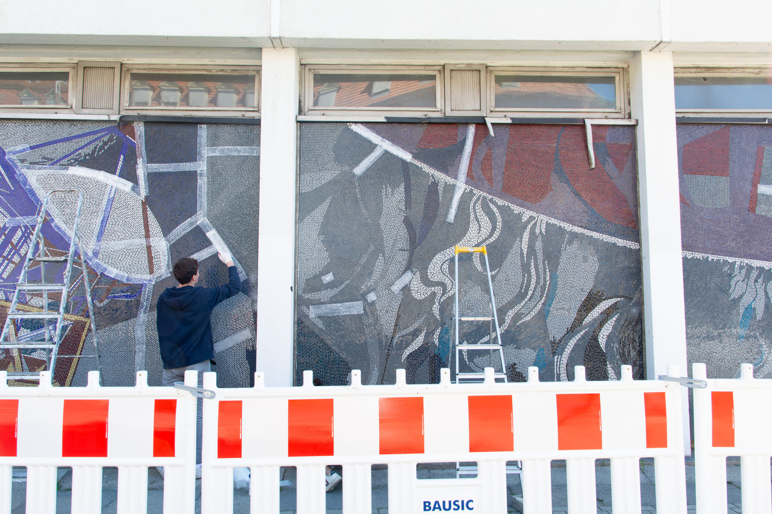 Hinter einem Bauzaun laufen die notdürftigen Restaurierungsmaßnahmen an den Mosaiktafeln. In diesem Bild sieht man, wie Japanpapier in Streifen aufgeklebt wird.