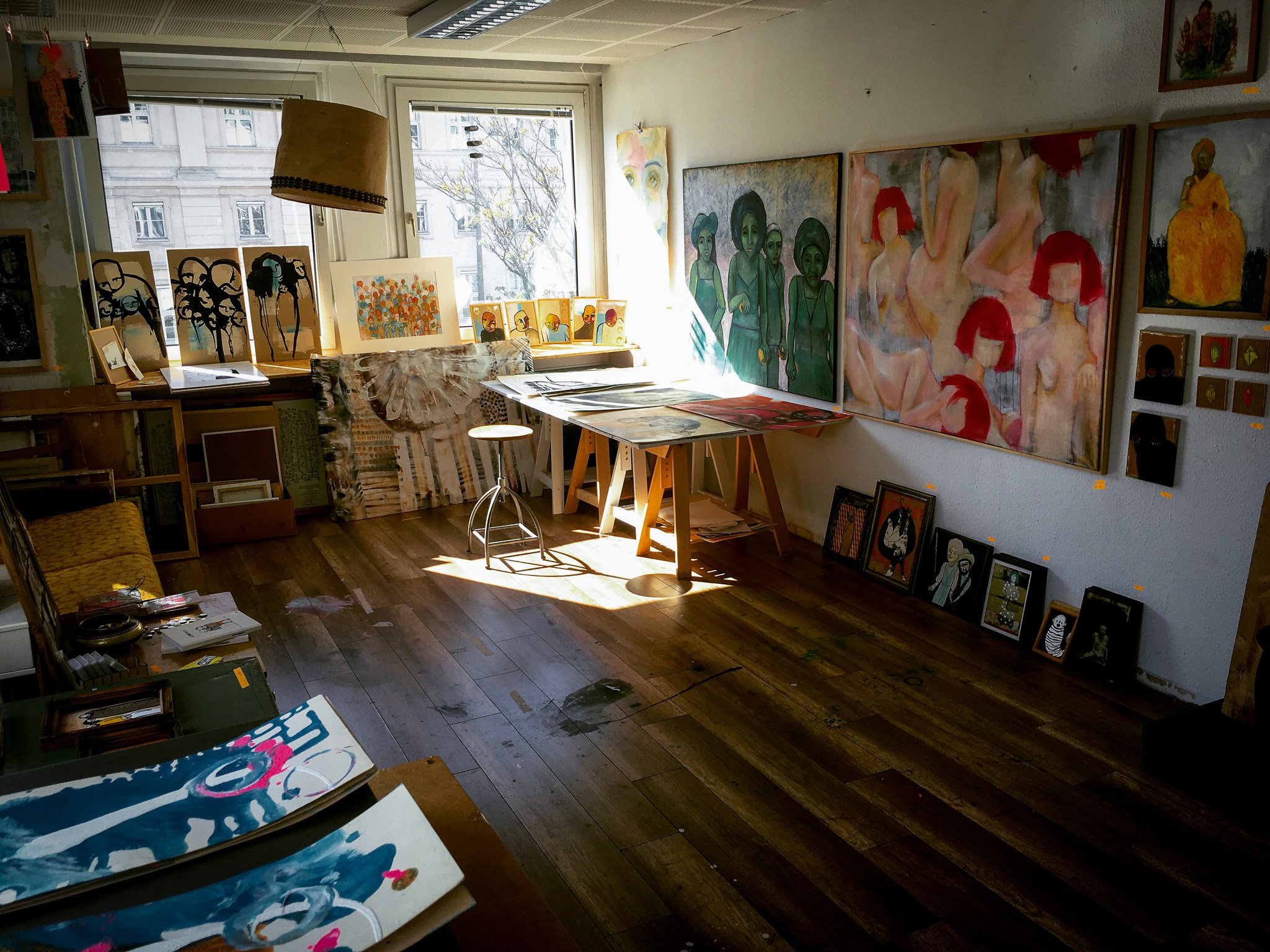 Ein Blick in ein Atelier: Auf einem Holzfußboden stehen verschiedene Gemälde, an den Wänden sind ebenfalls Gemälde mit Portraitdarstellungen.